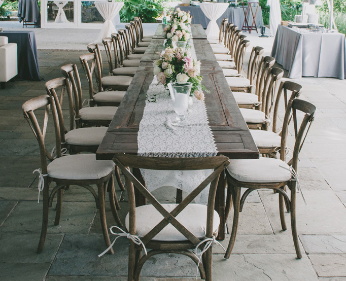 Természetes stílusú esküvői ülőgarnitúra kereszttámlás székekkel a teraszon, étkezőasztallal és virágdíszekkel